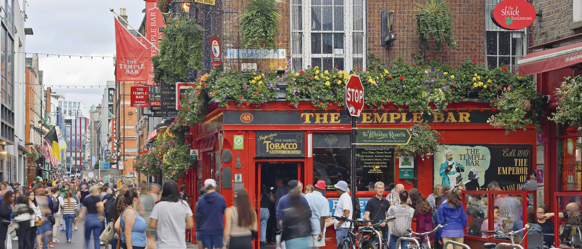 Busy street scene with people walking by the Temple Bar pub, adorned with greenery and colorful banners.