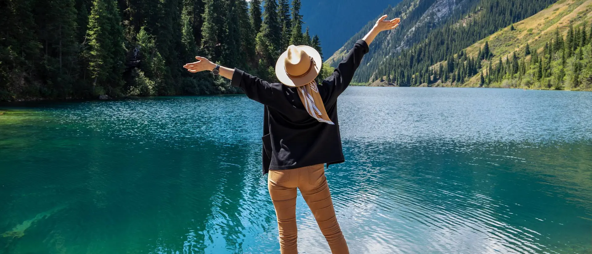 Person in a hat standing by a serene blue lake, arms outstretched, surrounded by trees and mountains under a clear sky.