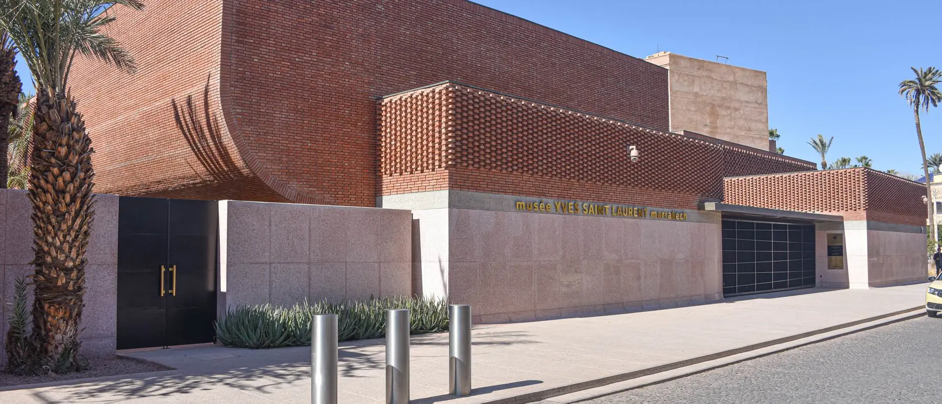 Modern brick building with palm trees and a paved walkway in front. Clear blue sky overhead.