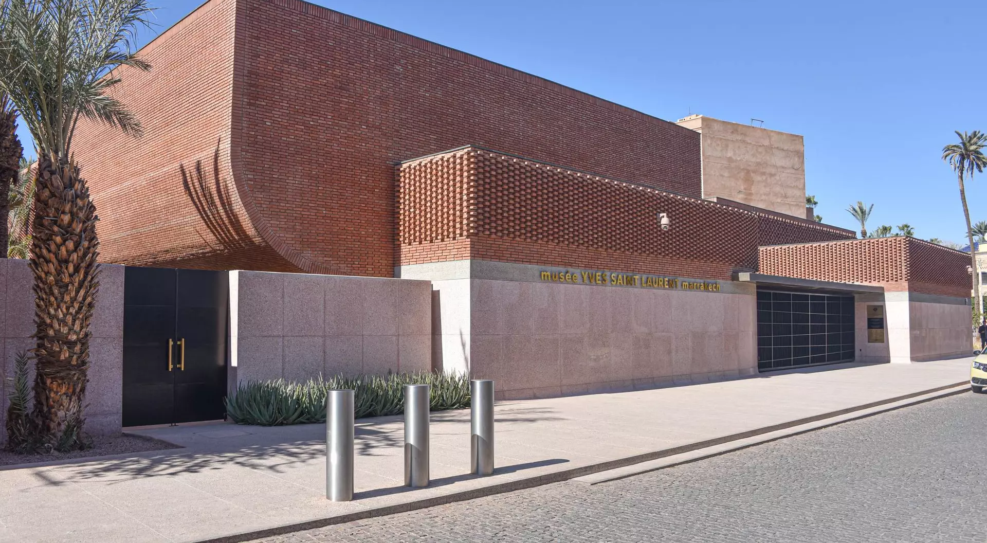 Modern brick building with palm trees and a paved walkway in front. Clear blue sky overhead.