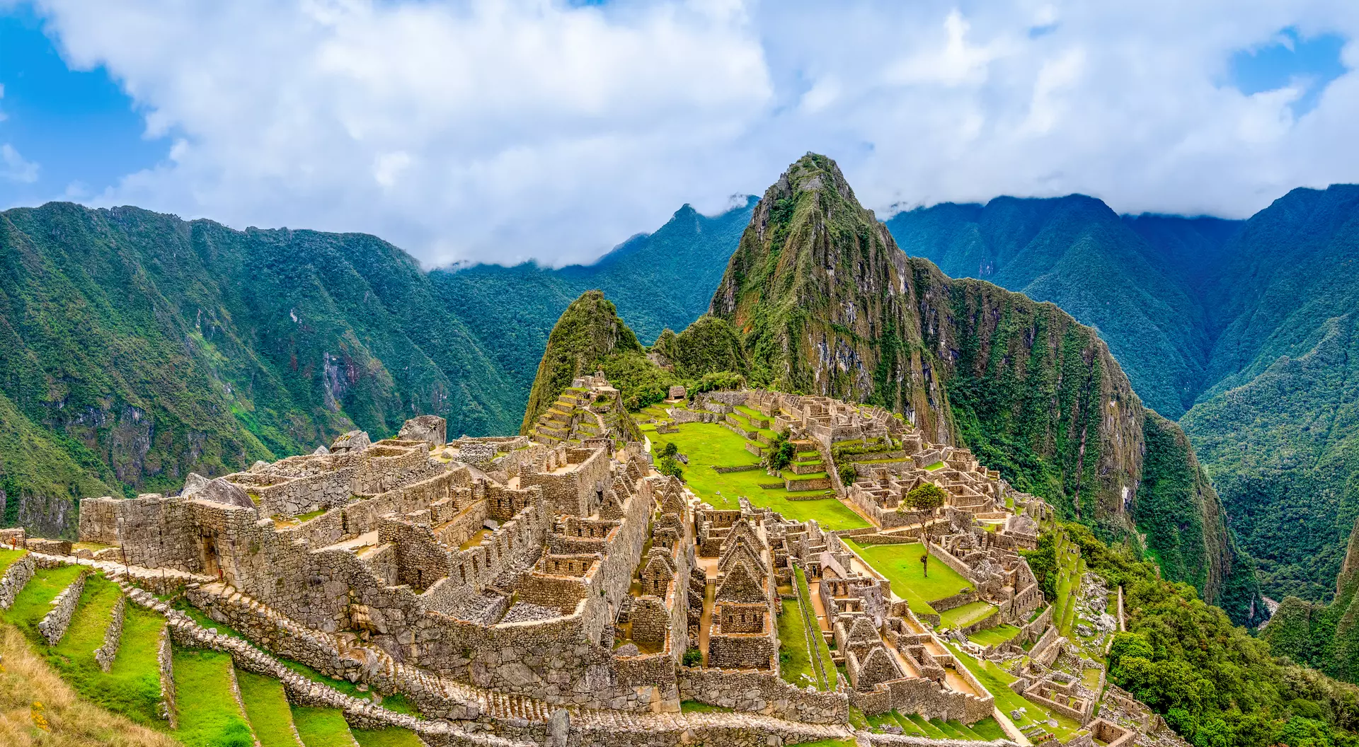 A panoramic view of Machu Picchu, an ancient Incan city, surrounded by mountains and lush greenery under a blue sky.