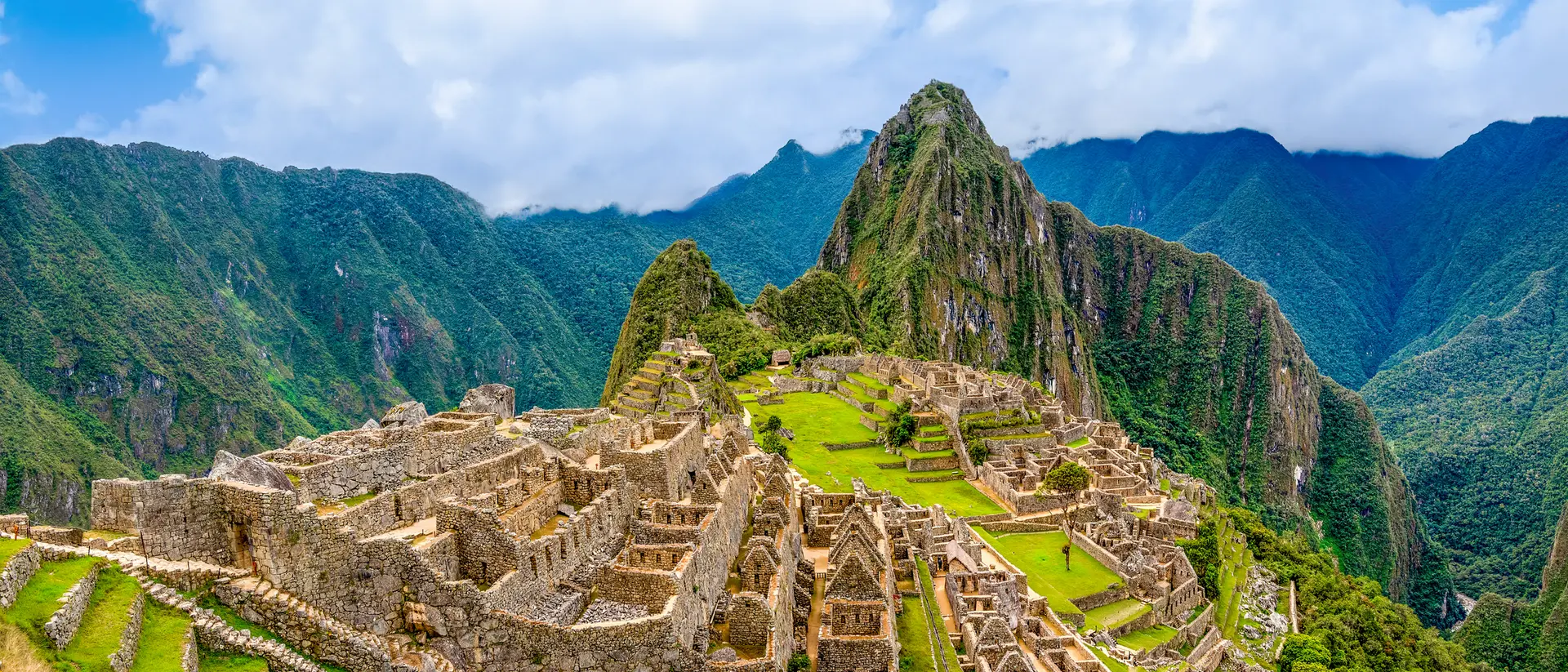 A panoramic view of Machu Picchu, an ancient Incan city, surrounded by mountains and lush greenery under a blue sky.
