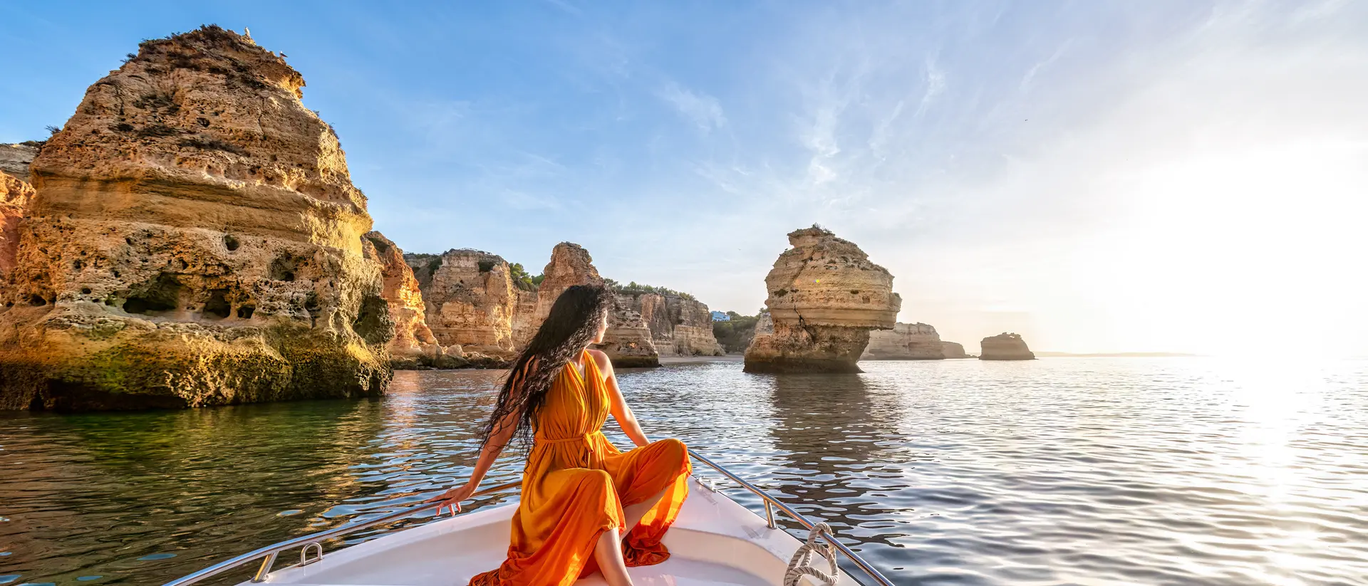 Person in a yellow dress sitting on the bow of a boat, gazing at rock formations and a sunset over calm water.