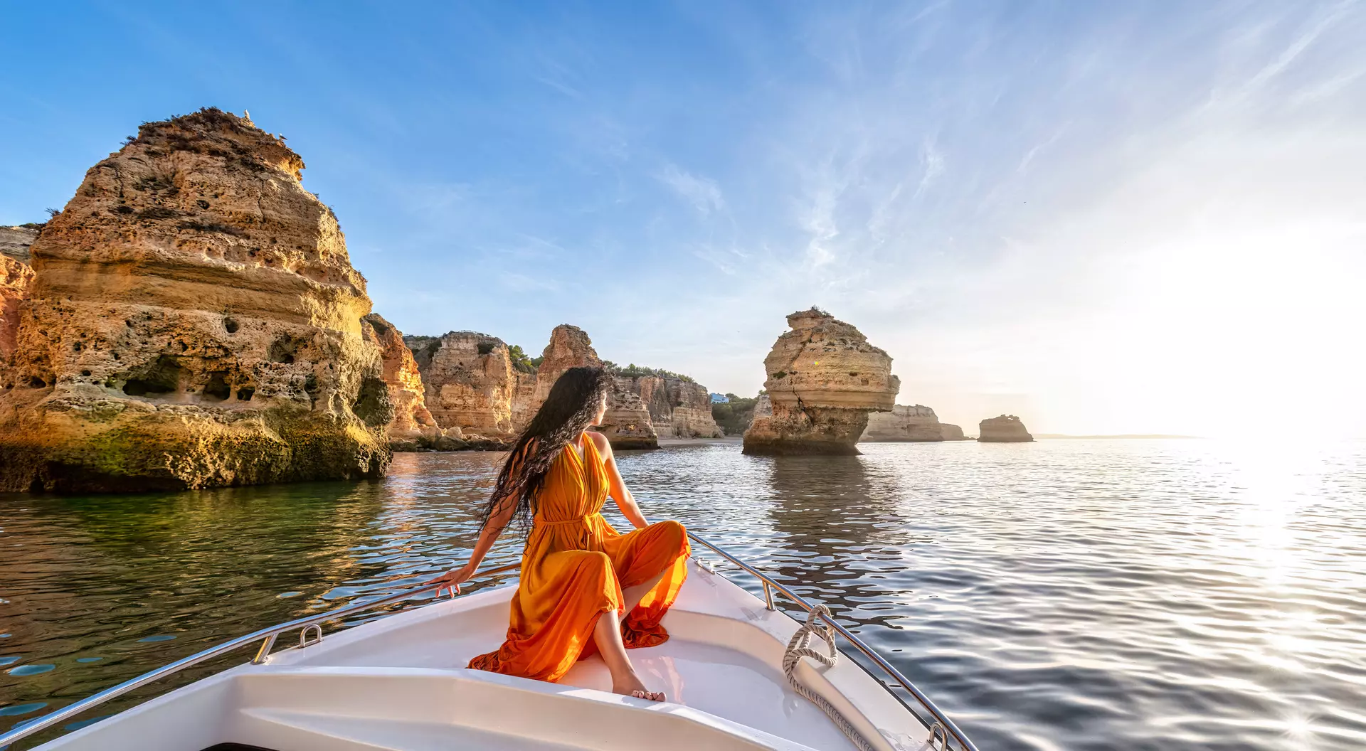 Person in a yellow dress sitting on the bow of a boat, gazing at rock formations and a sunset over calm water.