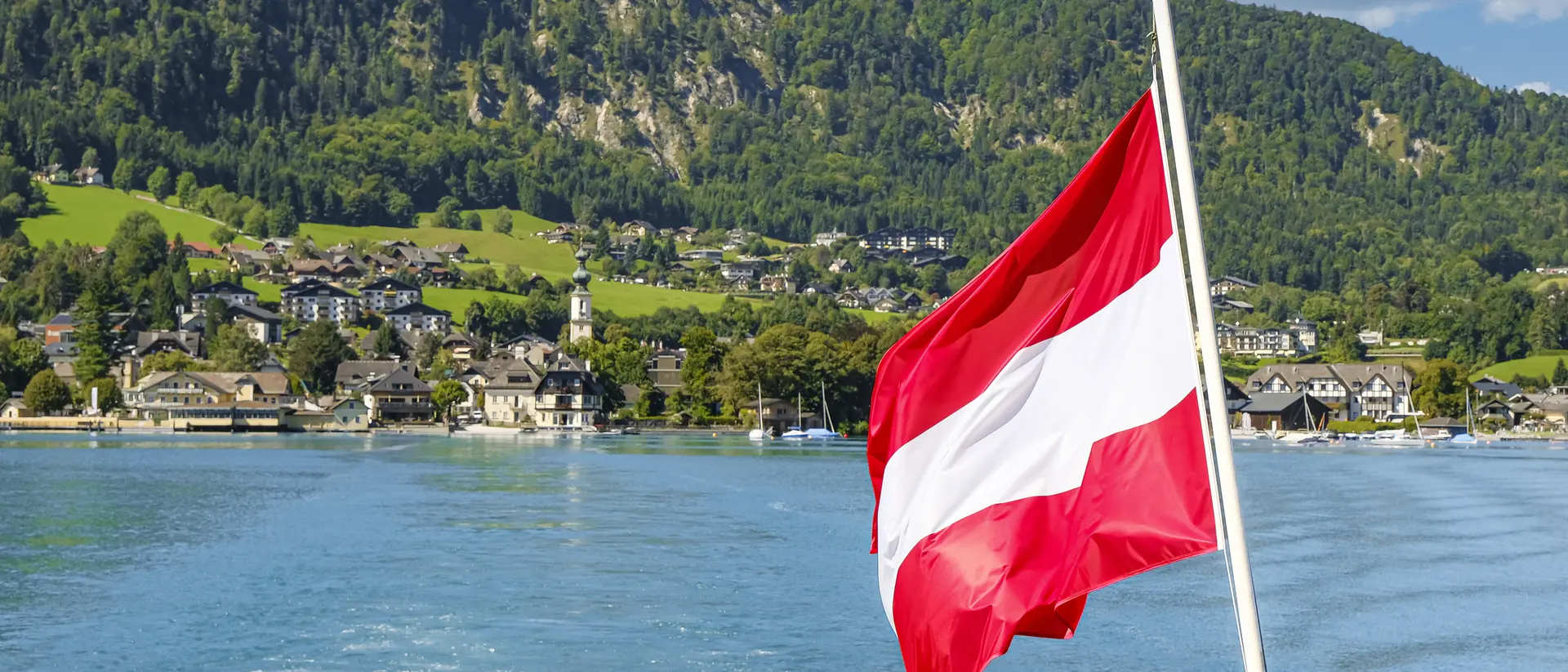 A red and white flag waves on a boat, overlooking a picturesque lakeside village surrounded by green mountains.