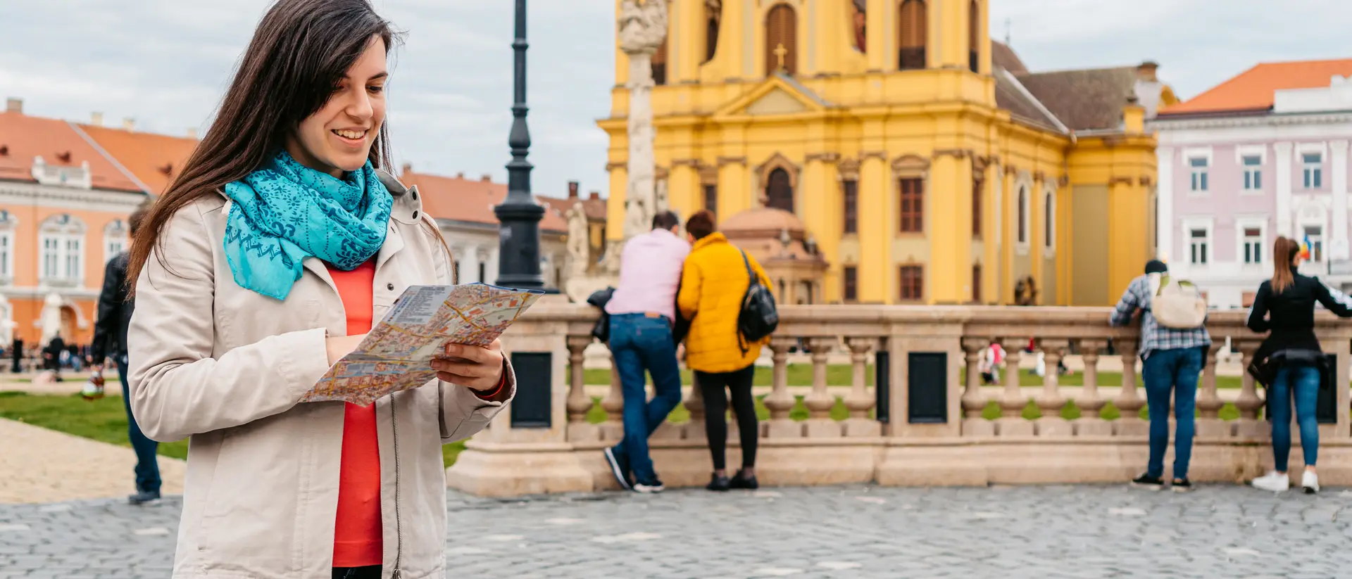 A woman with a map stands on a cobblestone street, colorful buildings and a yellow church visible in the background.