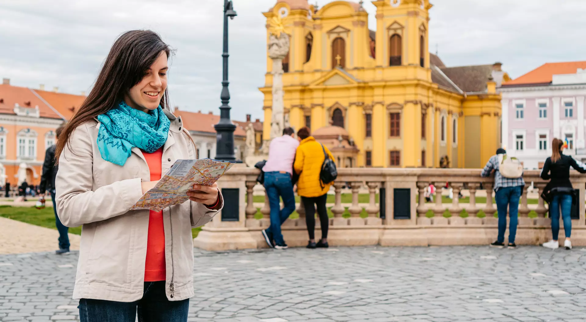 A woman with a map stands on a cobblestone street, colorful buildings and a yellow church visible in the background.