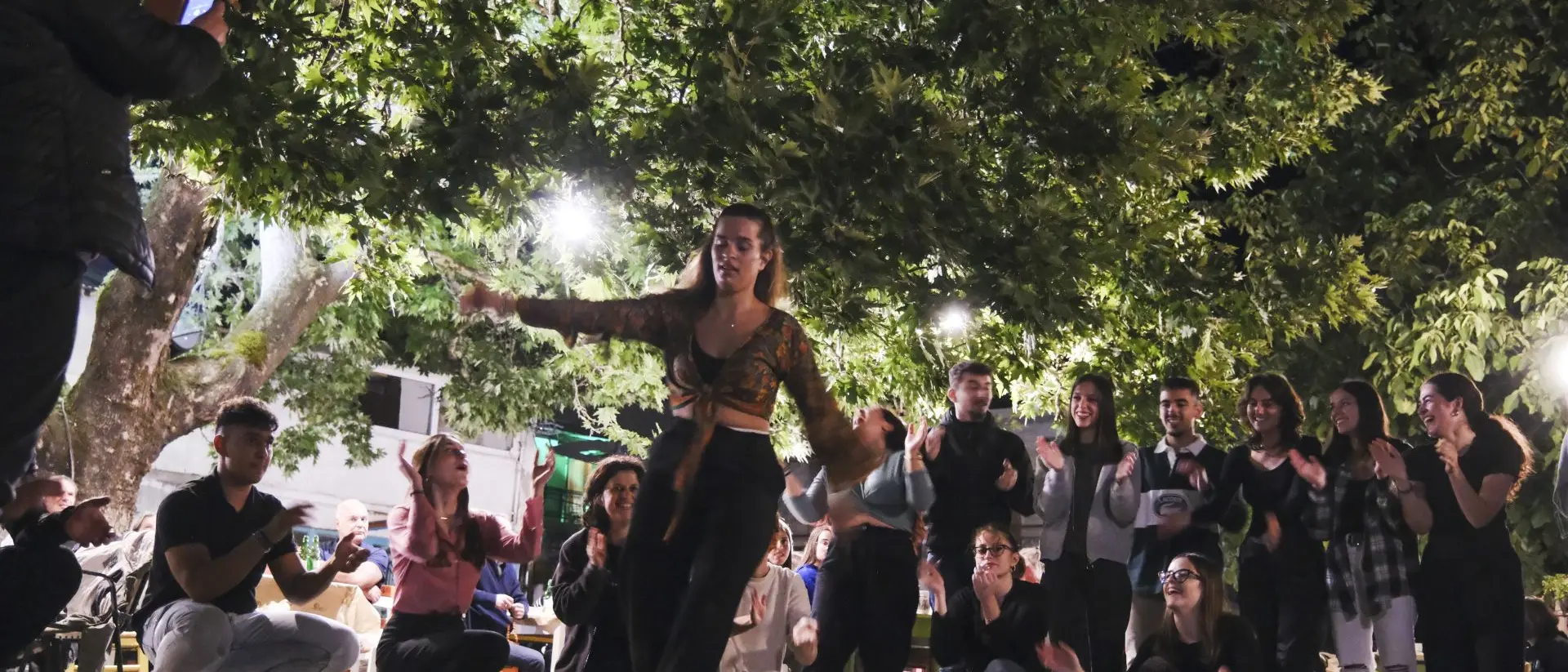 A lively outdoor scene with a dancer performing, surrounded by an engaged audience, under bright lights and trees.