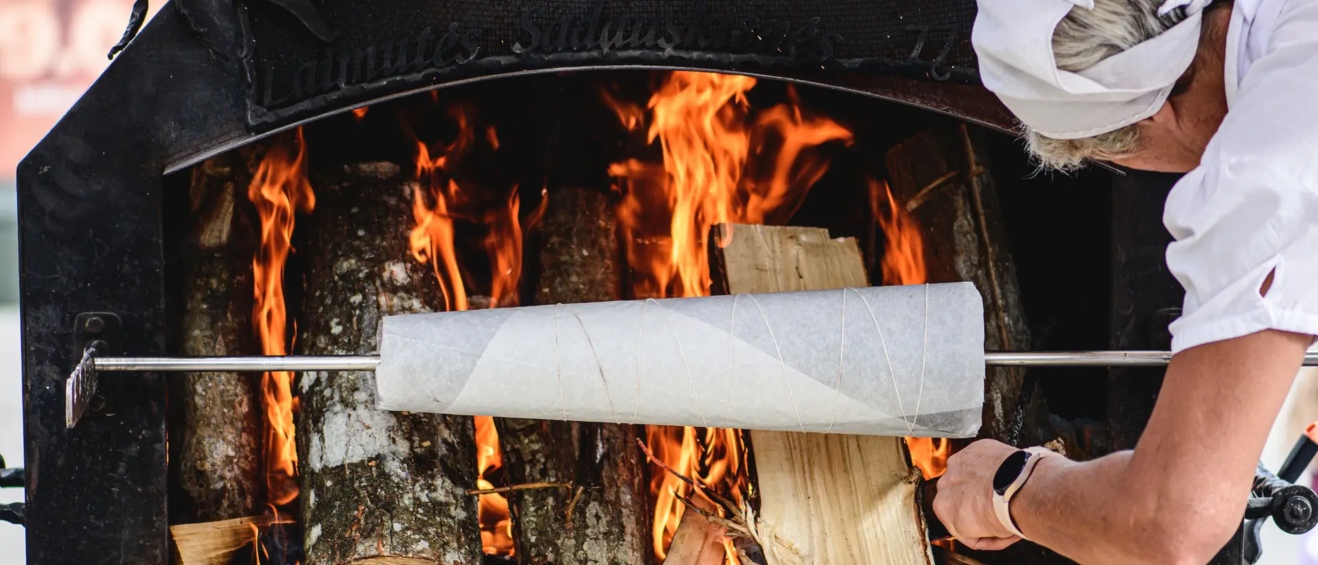 A person adjusts a cooking spit inside a wood-fired oven, with flames and logs visible in the background.