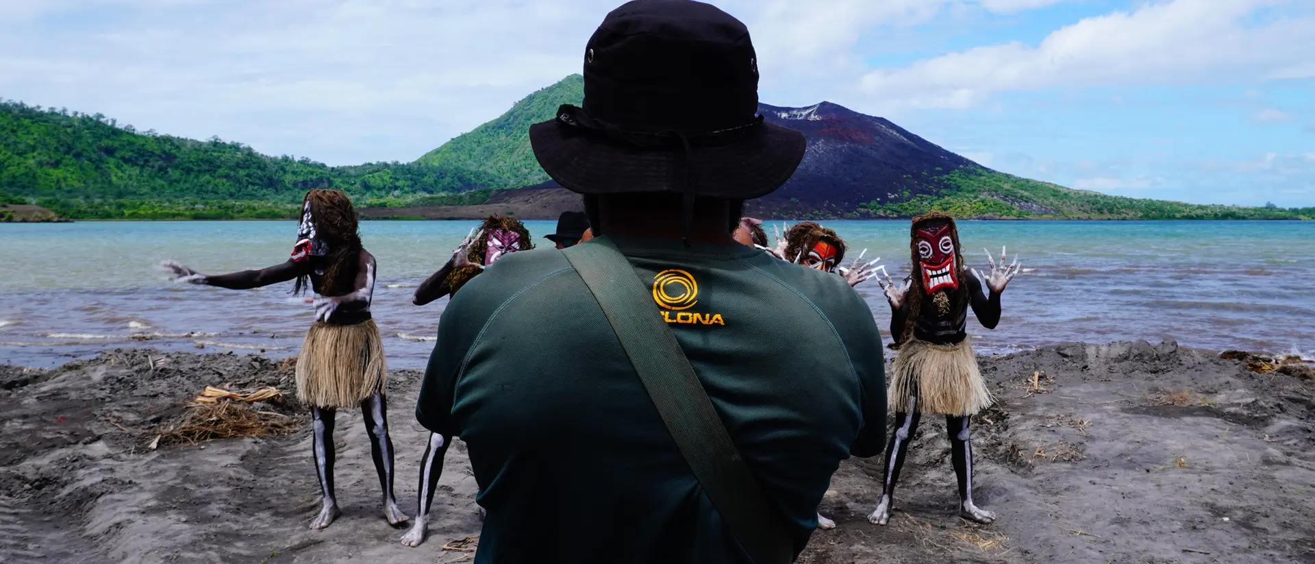 A person in a hat watches performers in masks and skirts dance by a serene waterfront and mountain backdrop.