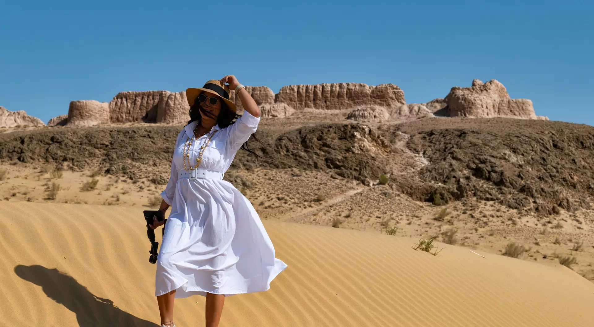 A person in a white dress and hat stands on sand dunes with desert cliffs in the background, holding a camera.