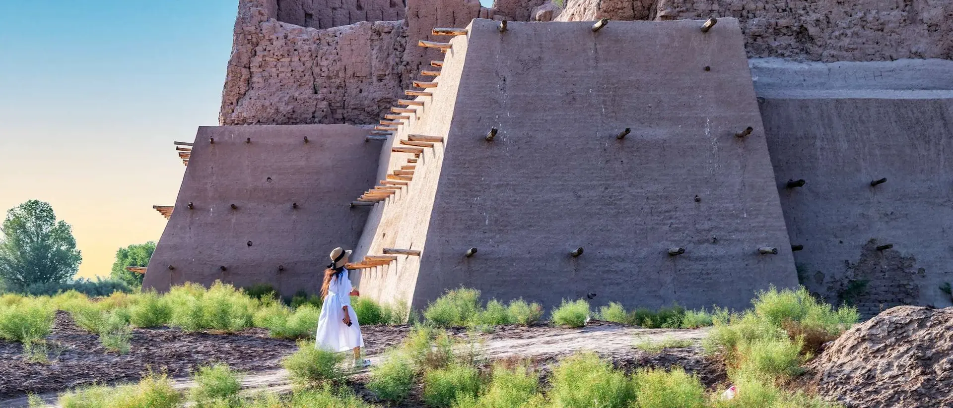 A person in a white dress stands near a large, ancient structure surrounded by green vegetation under a clear sky.