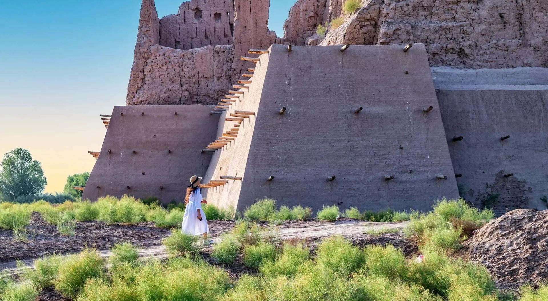 A person in a white dress stands near a large, ancient structure surrounded by green vegetation under a clear sky.