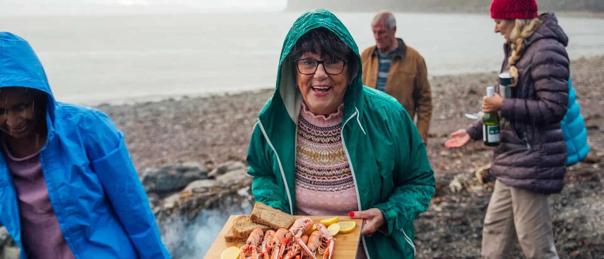 A smiling woman in a green jacket holds a tray of seafood by the beach, with friends nearby in casual attire.