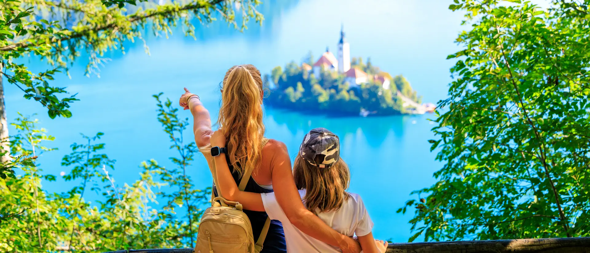 Two people stand by a railing, overlooking a serene blue lake with a small island and a church in the distance, surrounded by trees.
