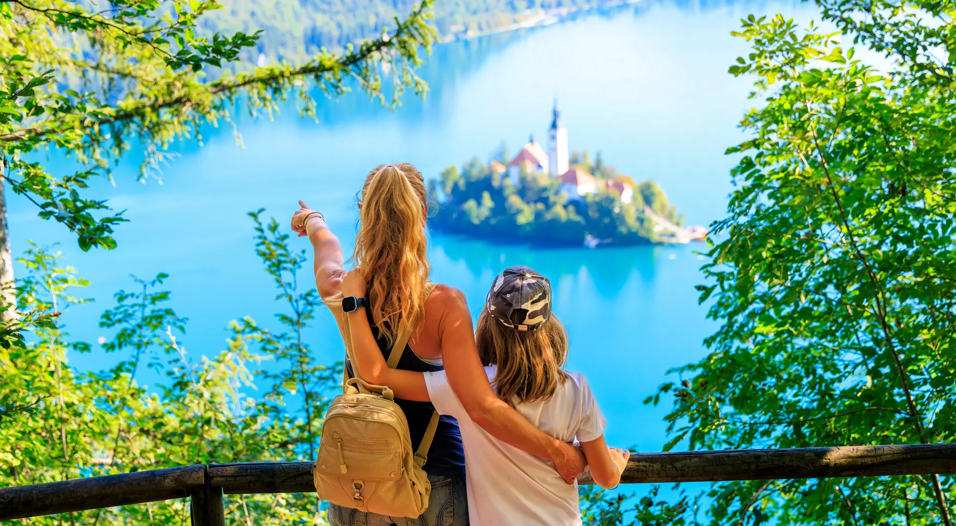 Two people stand by a railing, overlooking a serene blue lake with a small island and a church in the distance, surrounded by trees.
