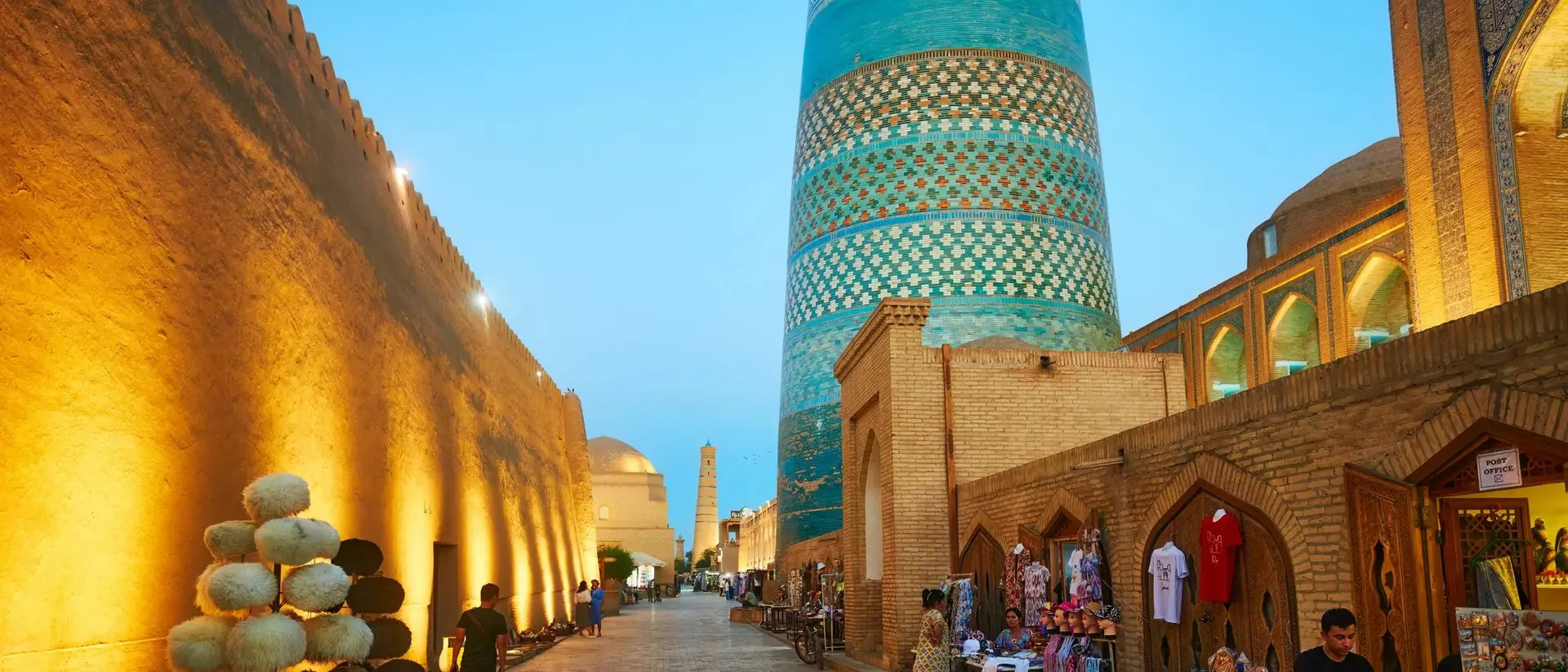 Evening scene in Khiva, Uzbekistan, showcasing a historic street lined with shops and a prominent blue minaret.