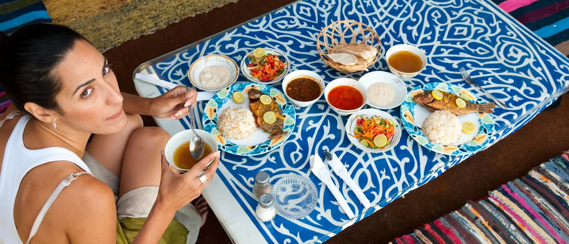 Person sitting on a colorful rug, enjoying a meal with plates of rice, fish, and various side dishes.