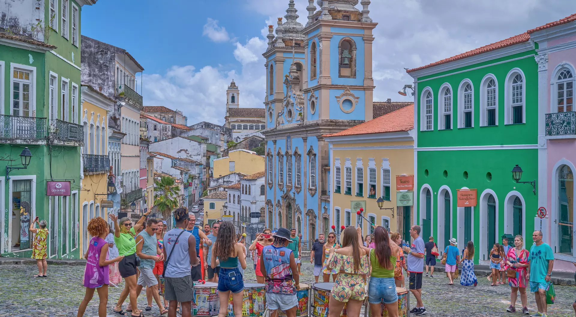 Colorful buildings line a cobblestone street while a group of people gathers around an art display under a cloudy sky.