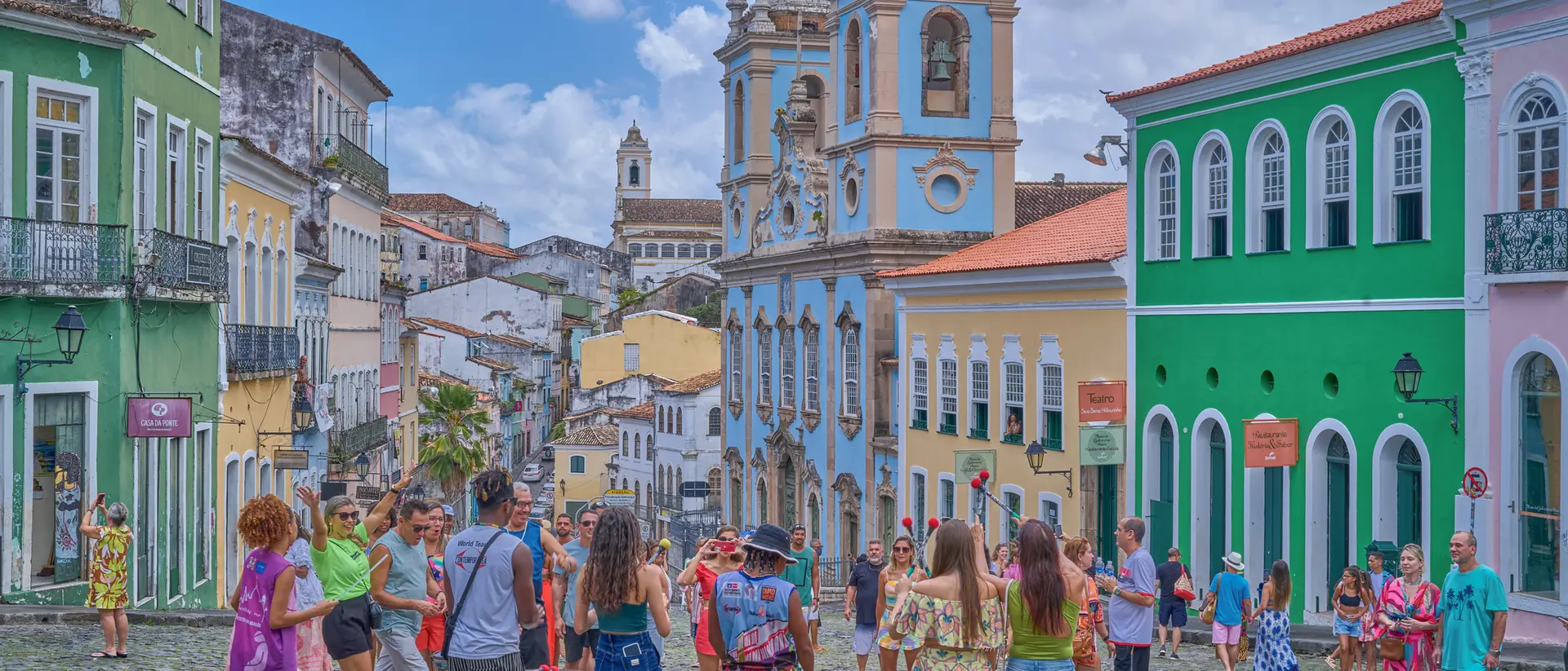 Colorful buildings line a cobblestone street while a group of people gathers around an art display under a cloudy sky.