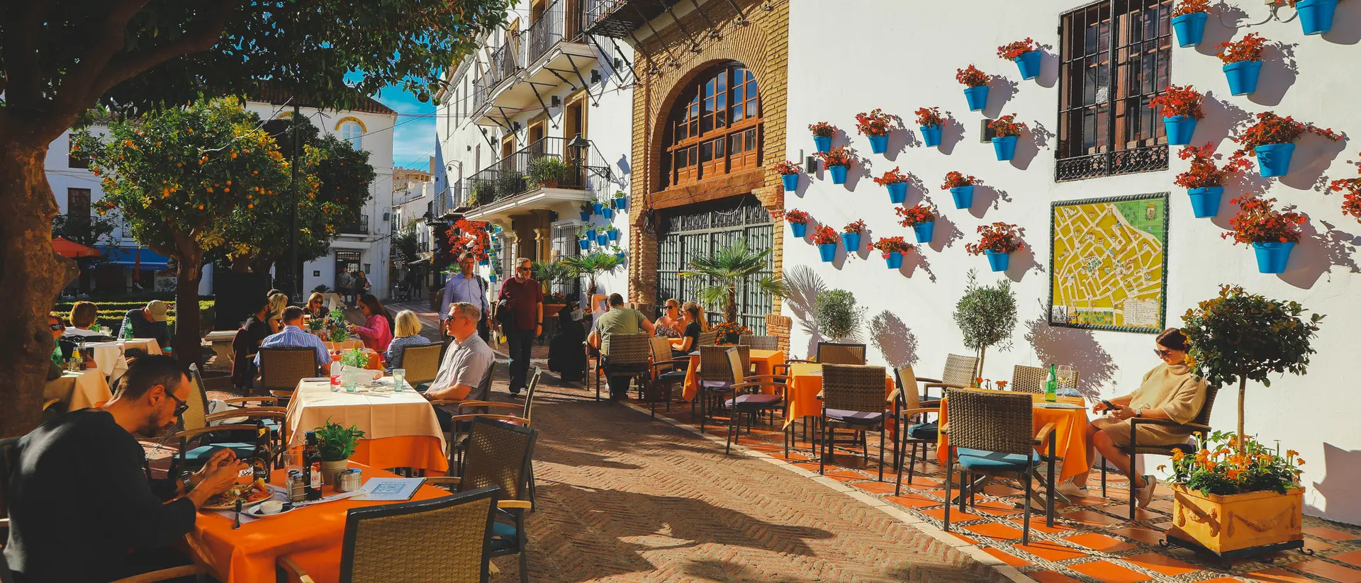 Outdoor café scene with people dining at tables, surrounded by vibrant flowers and a charming white building.
