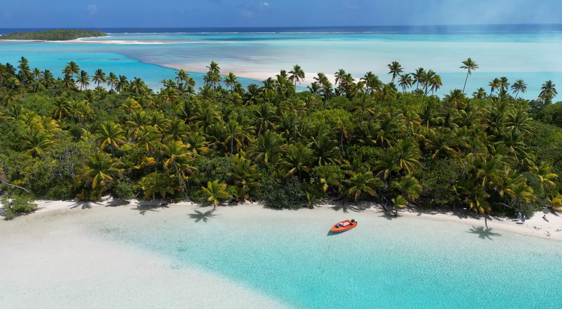 Aerial view of a tropical beach with turquoise water, a sandy shore, and lush green palm trees. A small boat is visible.