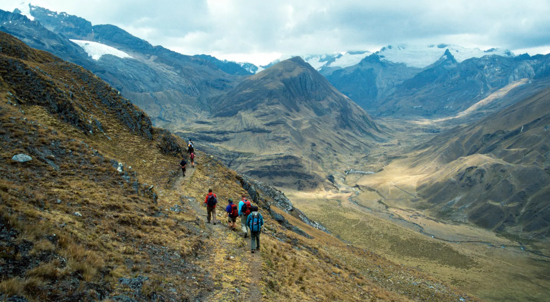 Hikers trek along a mountain trail overlooking a vast valley with snow-capped peaks in the distance.