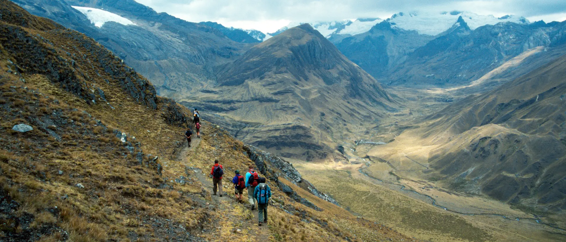 Hikers trek along a mountain trail overlooking a vast valley with snow-capped peaks in the distance.