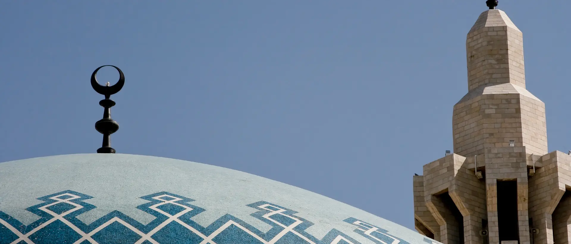Close-up of a mosque dome with geometric patterns, featuring a crescent moon and a tall minaret in the background.