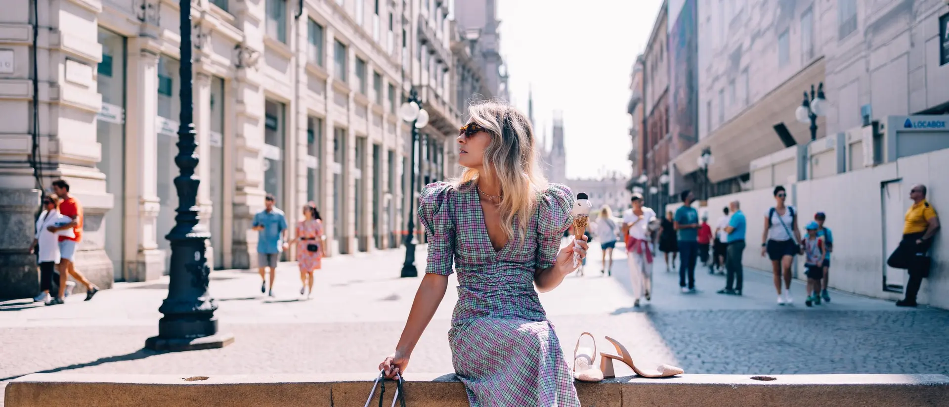 A woman in a patterned dress sits on a ledge, holding shopping bags, while people stroll by in a busy city street.
