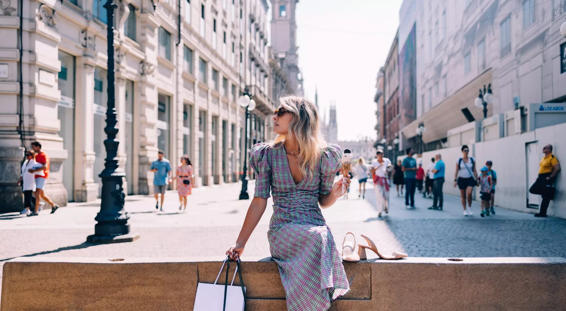 A woman in a patterned dress sits on a ledge, holding shopping bags, while people stroll by in a busy city street.