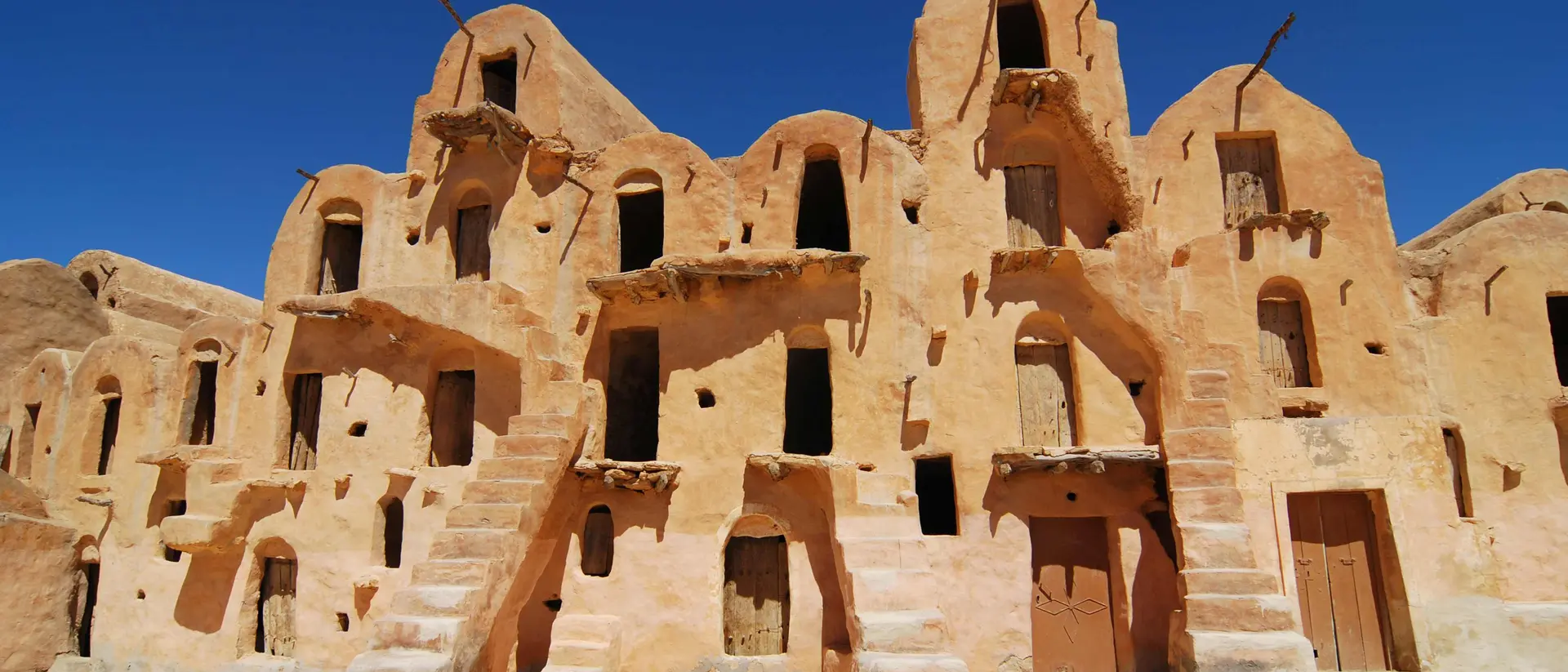 Ancient desert architecture featuring multi-level, ochre-colored buildings with stairs and numerous windows under a blue sky.