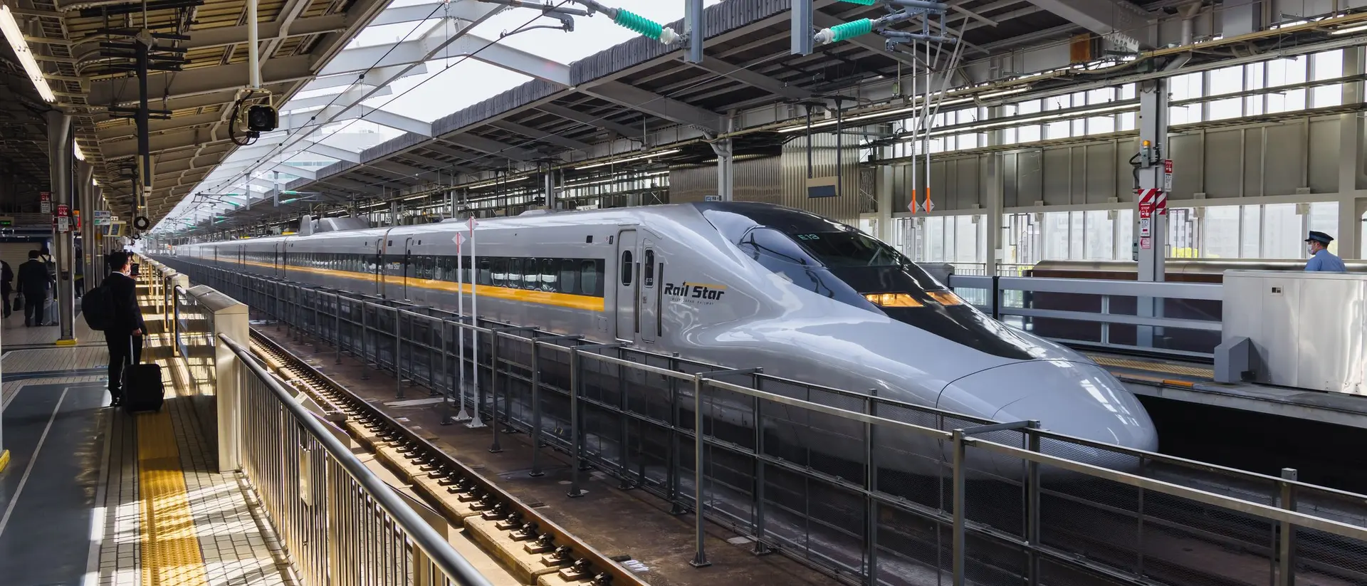 A sleek Shinkansen bullet train waits at a modern train station, with bright sunlight streaming through the platform.