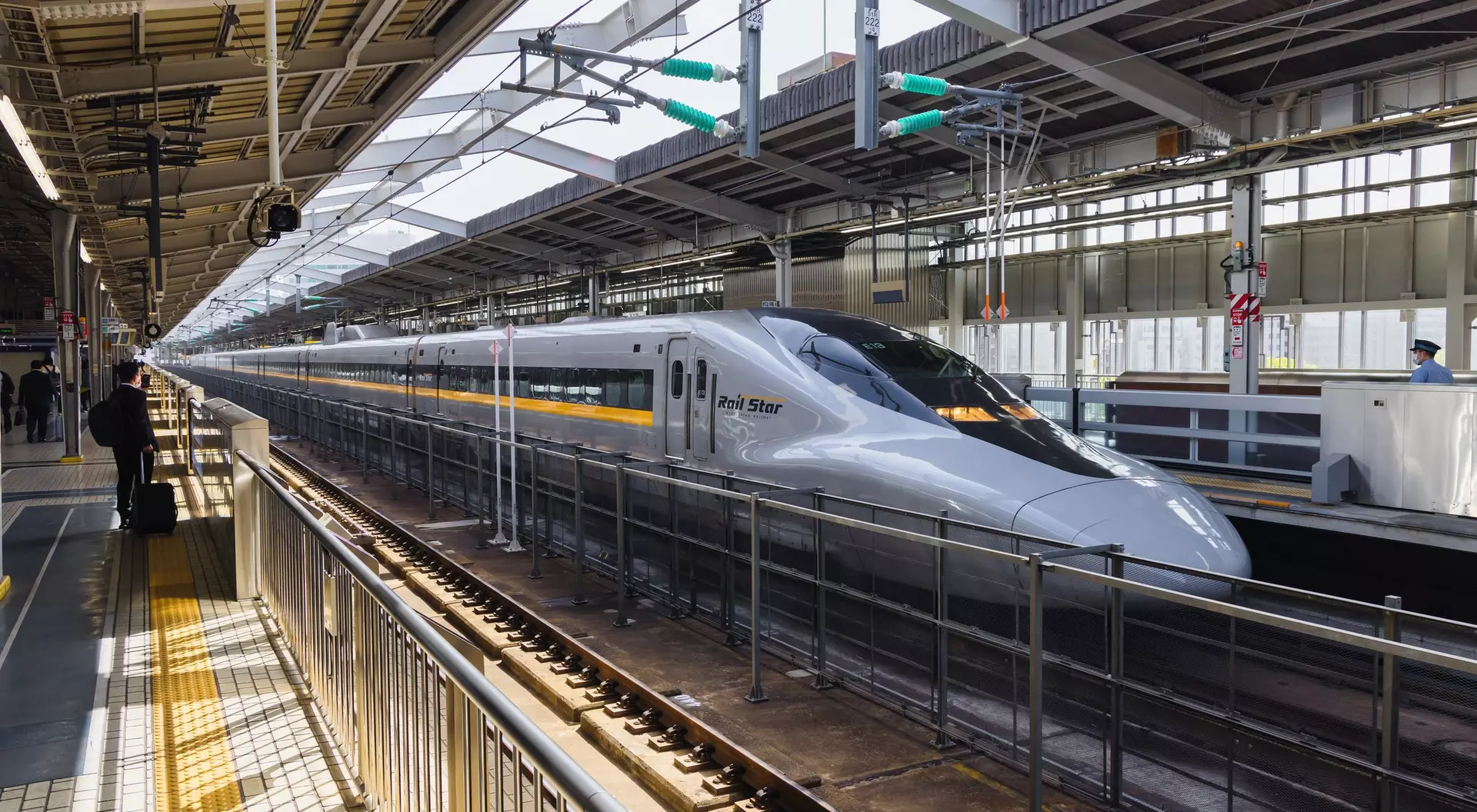 A sleek Shinkansen bullet train waits at a modern train station, with bright sunlight streaming through the platform.