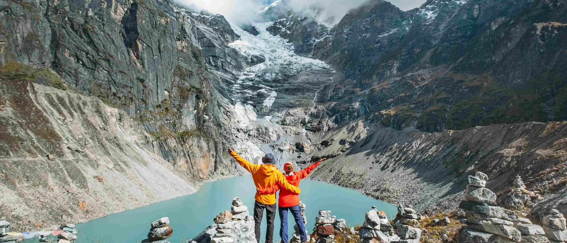 Two hikers in bright orange jackets stand atop a rocky overlook, celebrating the stunning mountain landscape and glacial lake below.