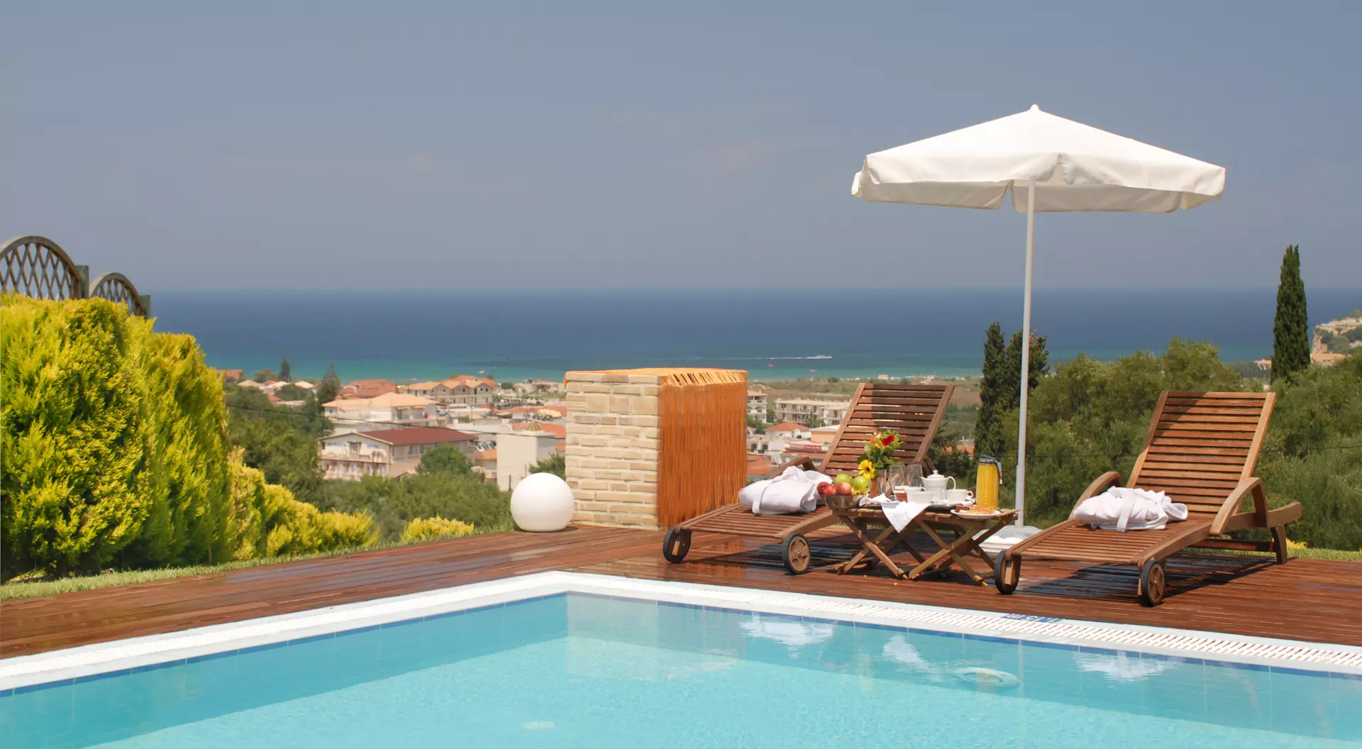 A sunny poolside scene with two lounge chairs and an umbrella overlooking a coastal view and lush greenery.