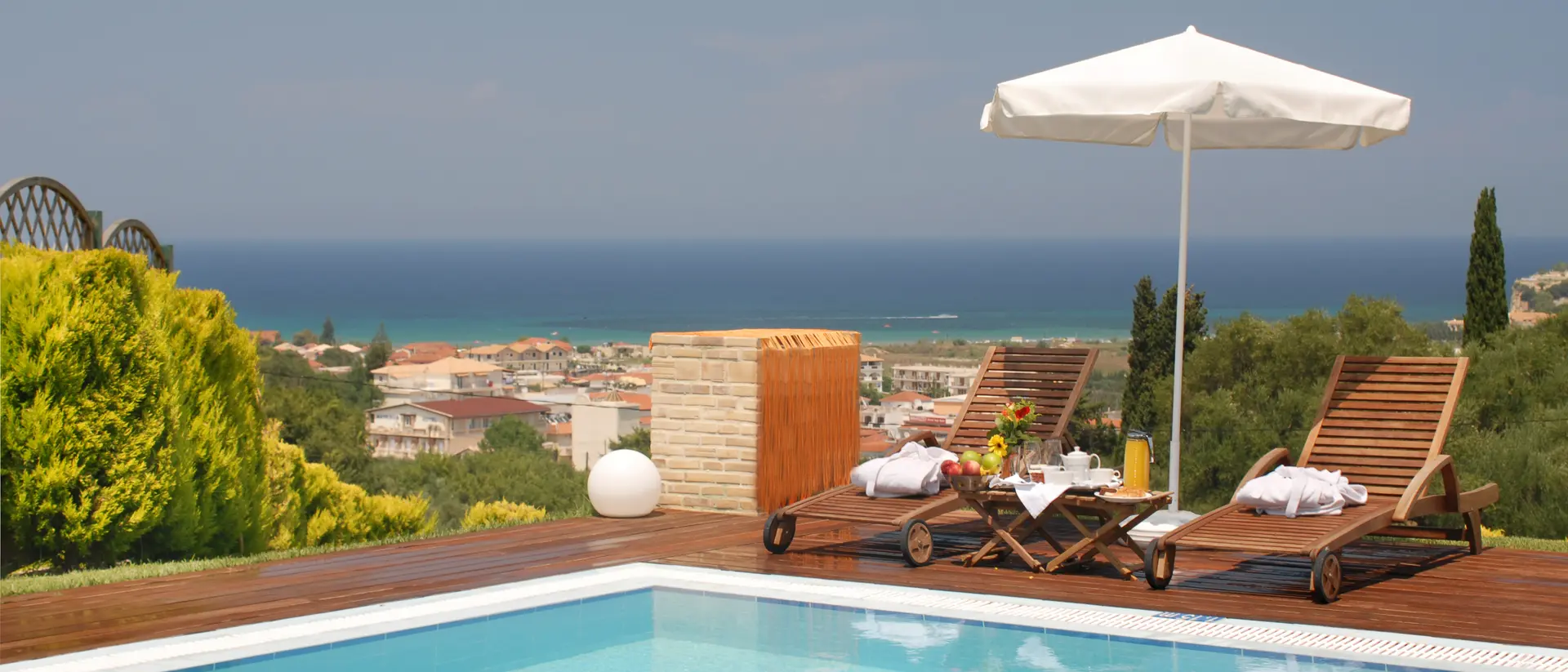 A sunny poolside scene with two lounge chairs and an umbrella overlooking a coastal view and lush greenery.