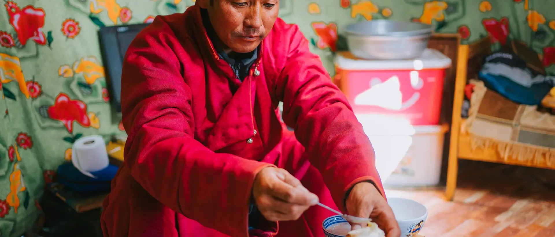 A man in a red robe serves food from a plate while sitting indoors, surrounded by colorful patterned walls.