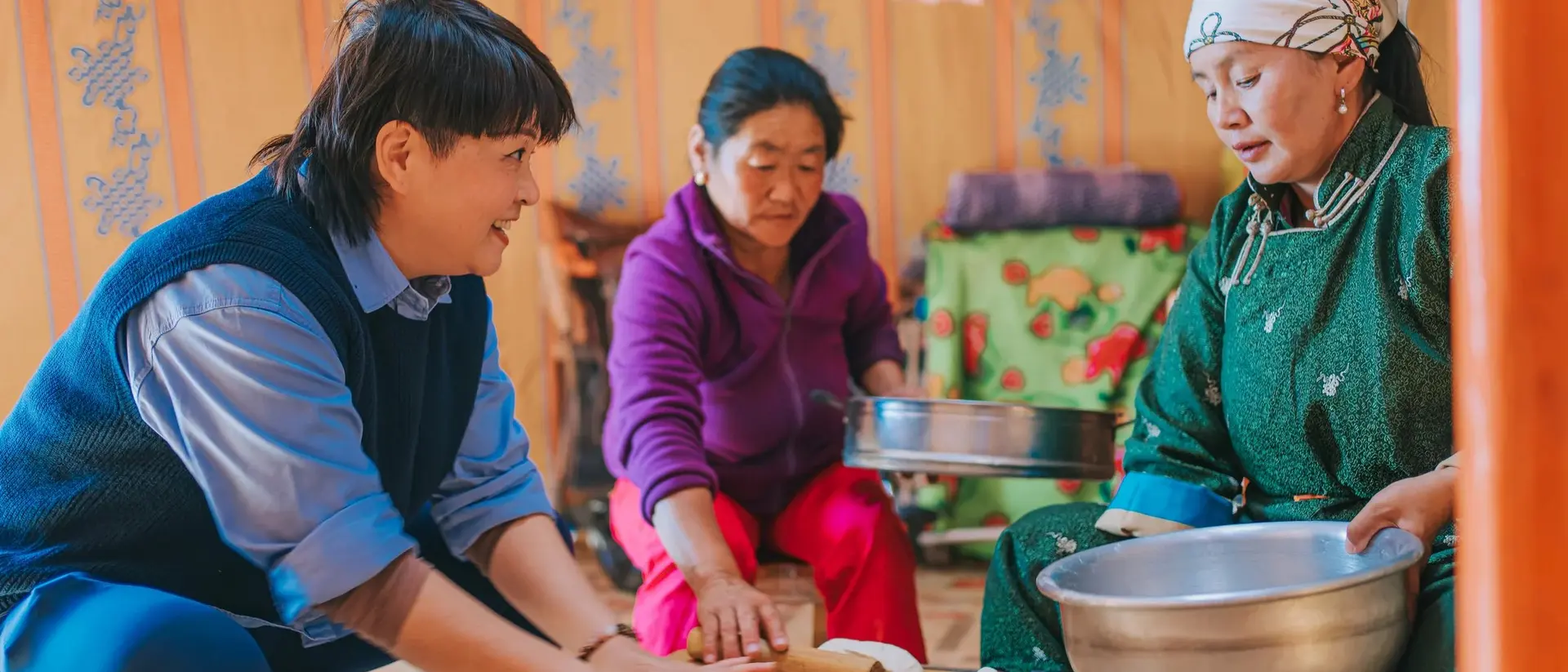 Three people prepare food inside a traditional yurt, with one person rolling dough and the others assisting at a table.