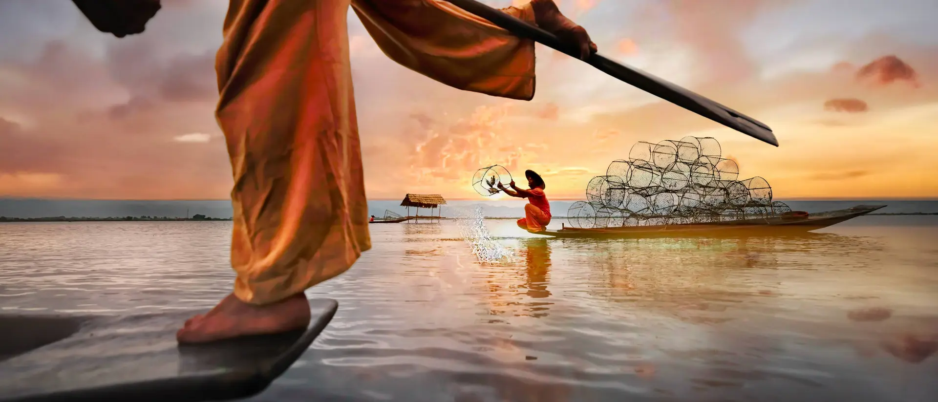 A fisherman in traditional dress uses a spear, while a woman in a boat casts nets at sunset over calm water.