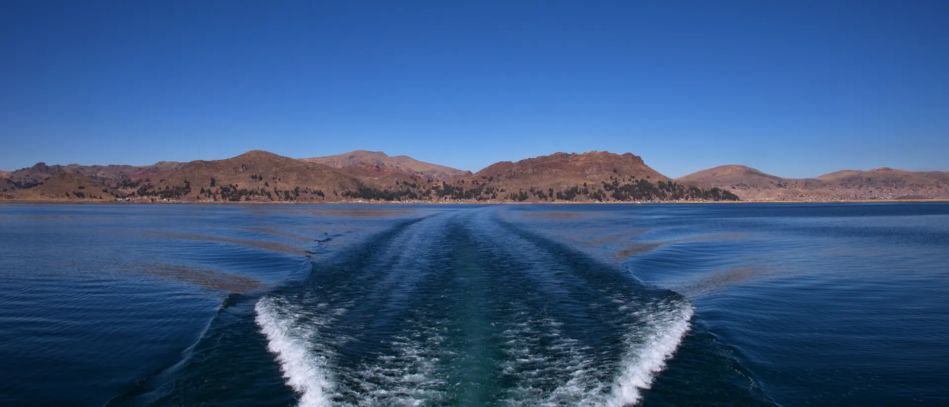 Wake left by a boat on a calm lake, with mountains and clear blue sky in the background.
