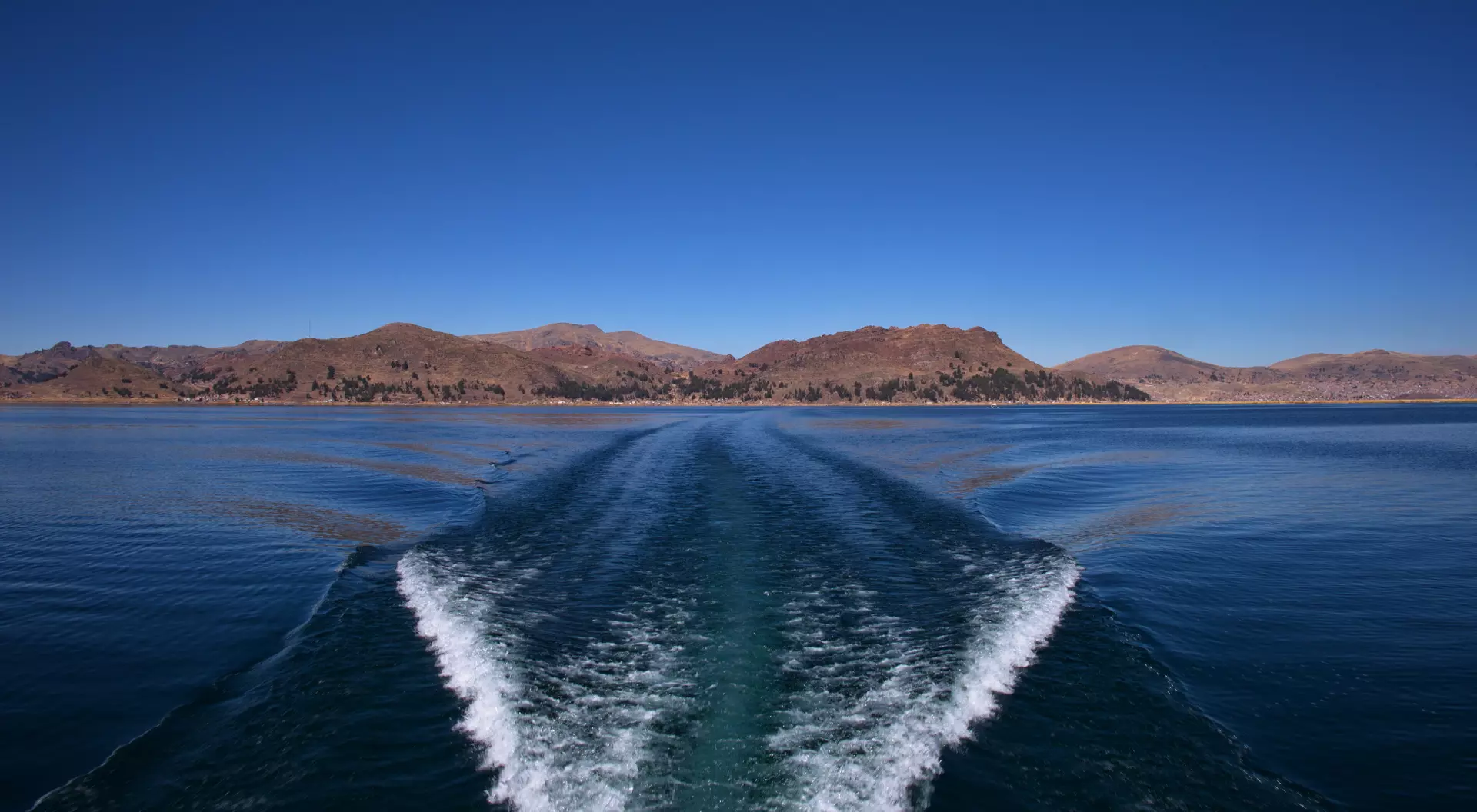 Wake left by a boat on a calm lake, with mountains and clear blue sky in the background.