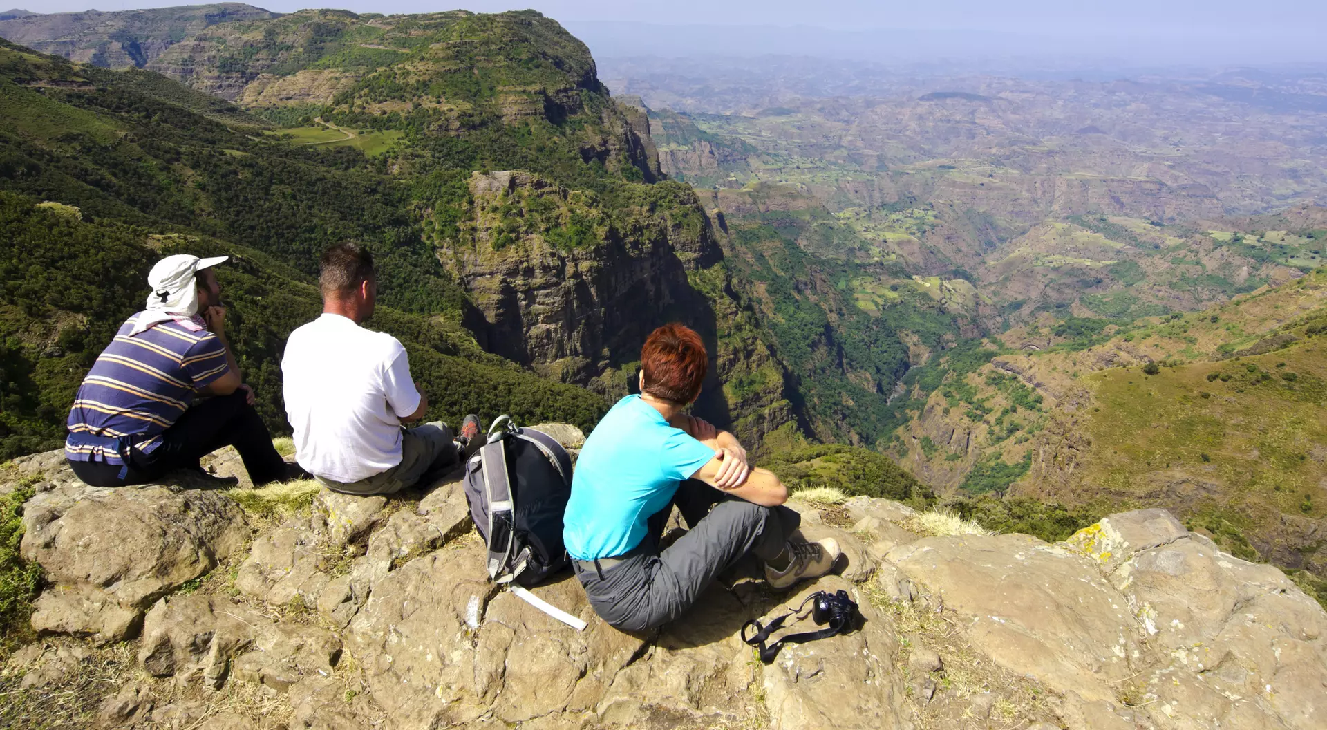Three people sit on a rocky ledge, overlooking a vast mountainous landscape under a clear blue sky.