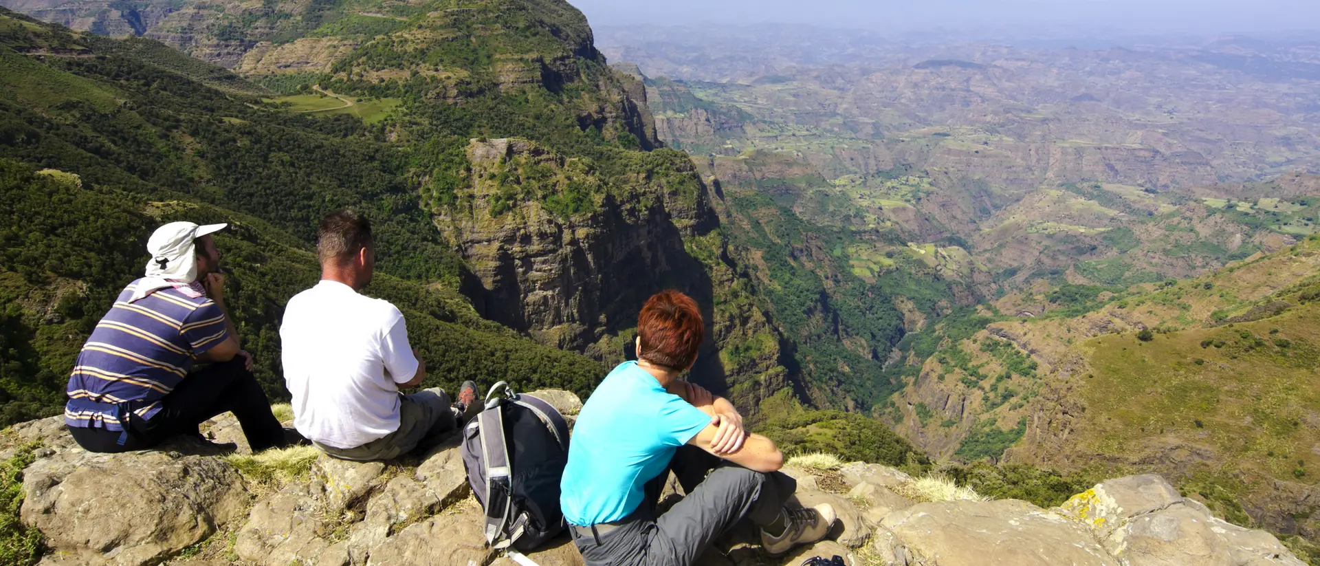 Three people sit on a rocky ledge, overlooking a vast mountainous landscape under a clear blue sky.