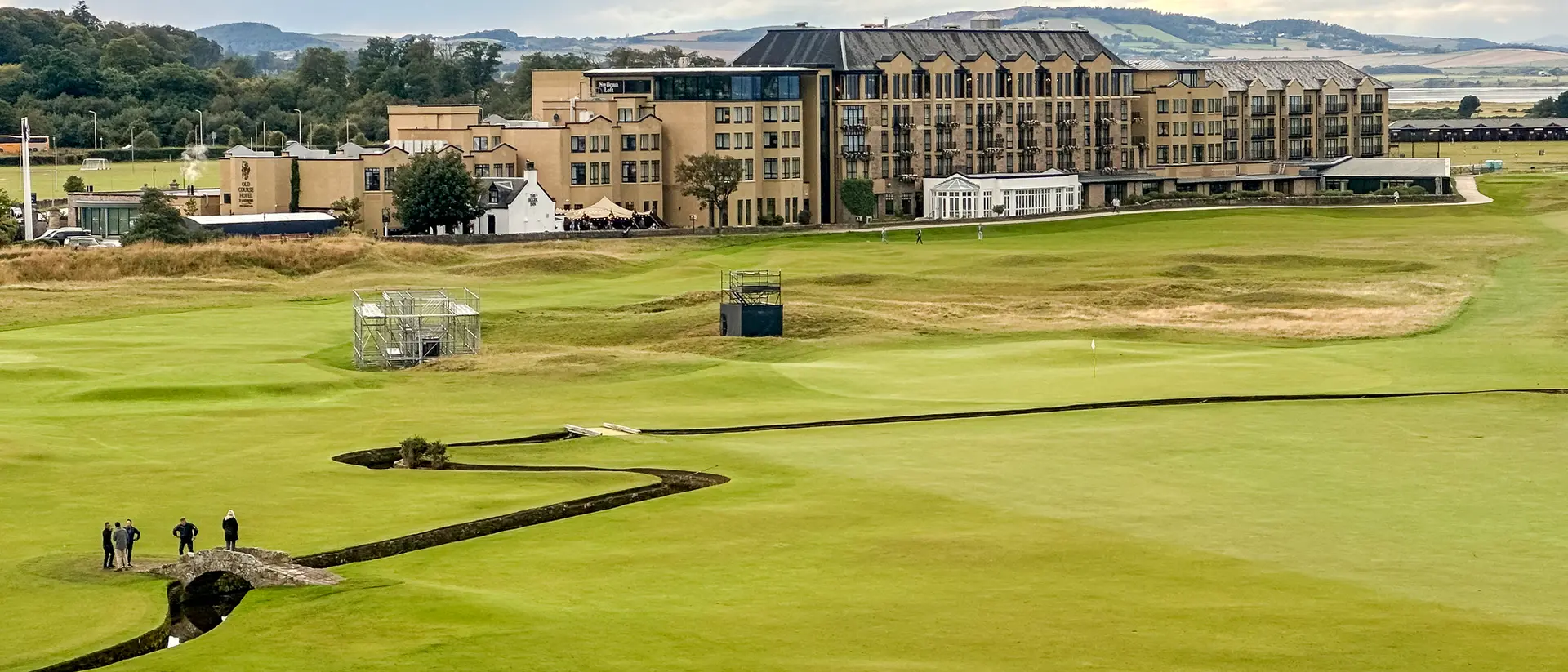 Landscape of a golf course with a hotel in the background, under a cloudy sky and a group of people on the green.