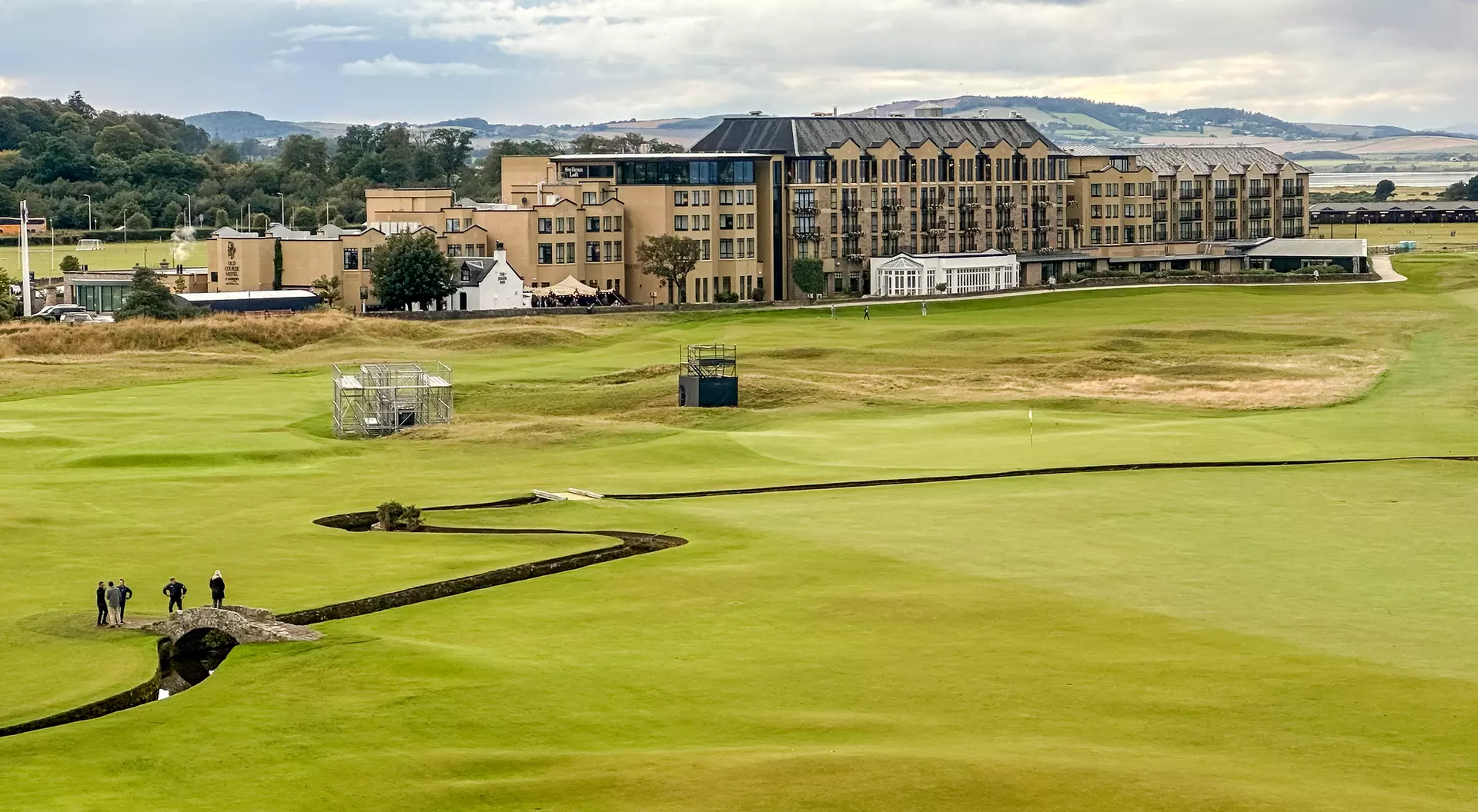 Landscape of a golf course with a hotel in the background, under a cloudy sky and a group of people on the green.