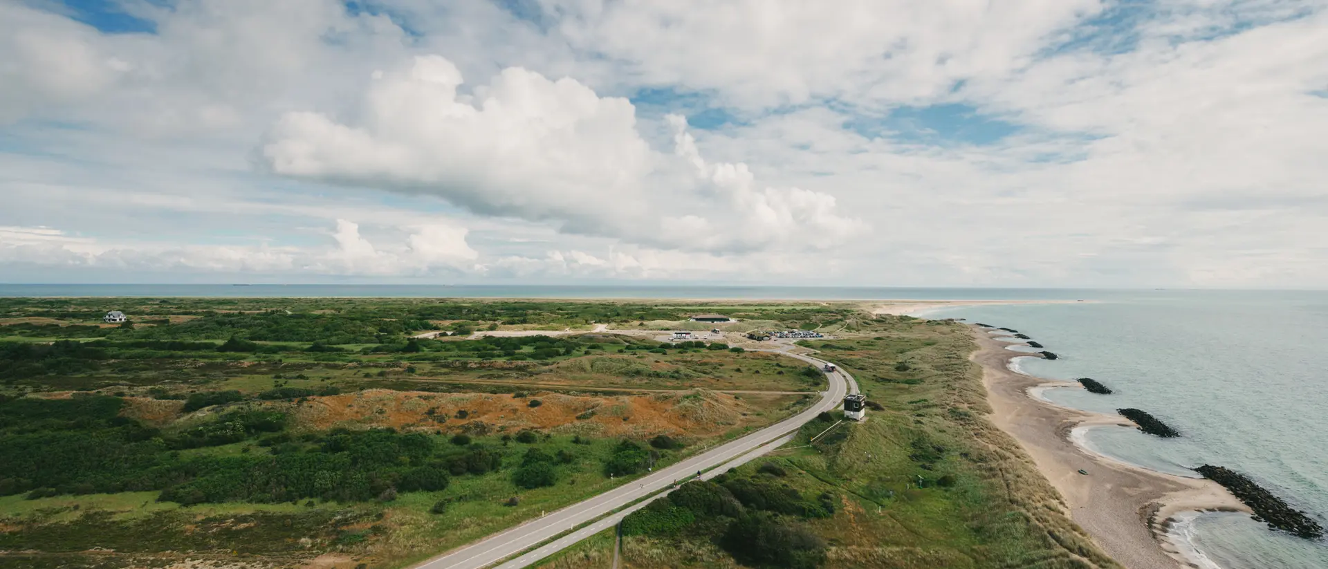 Aerial view of a coastal landscape with a winding road, green land, and ocean under a blue sky with clouds.