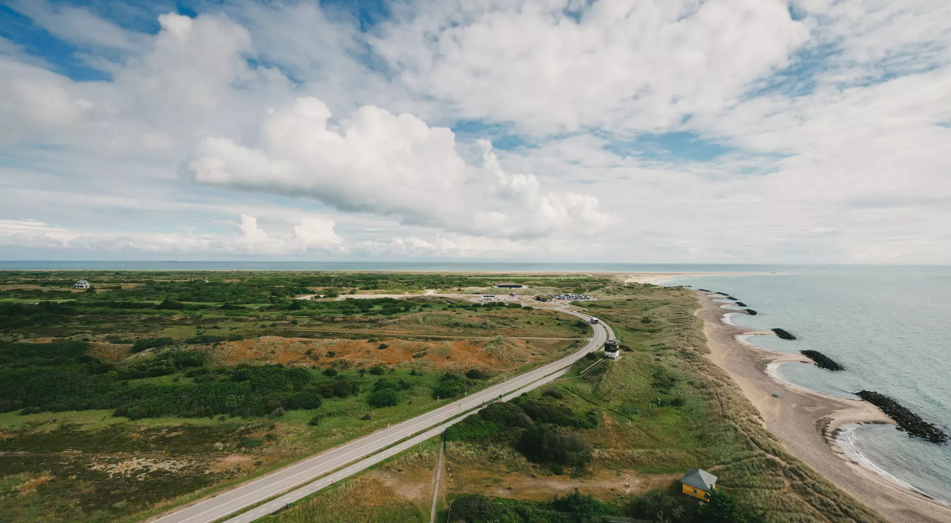 Aerial view of a coastal landscape with a winding road, green land, and ocean under a blue sky with clouds.
