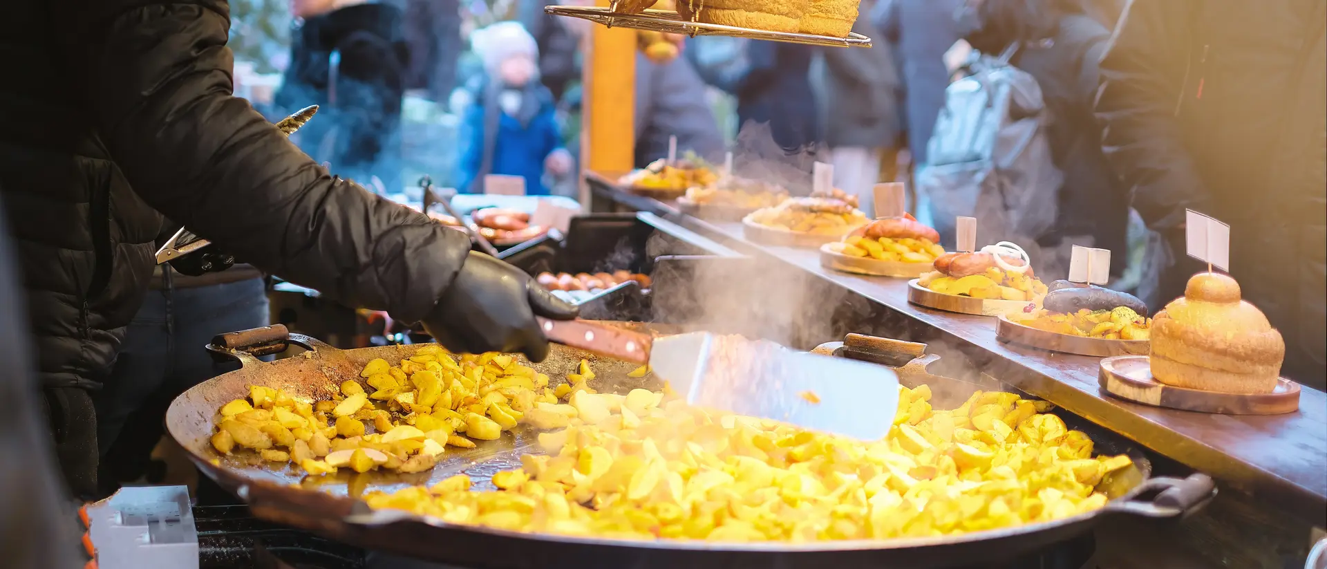 A vendor stirs a large pan of golden potatoes at a bustling outdoor market, surrounded by people and food stalls.