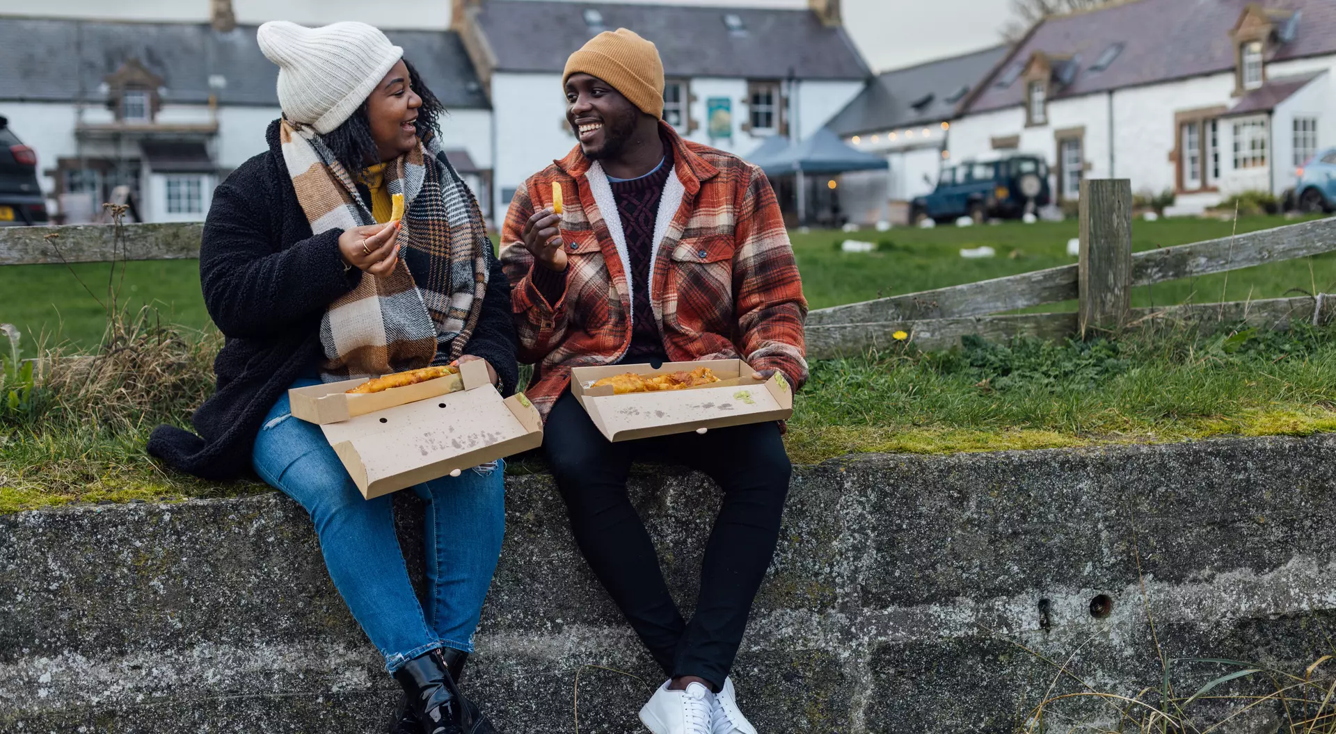 Two people enjoy food from takeout boxes while sitting on a stone wall, with buildings and greenery in the background.